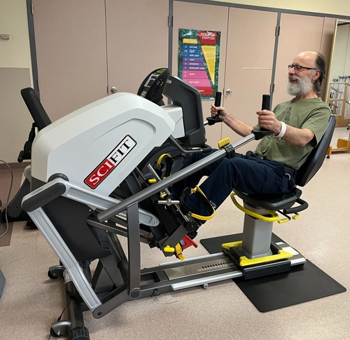 A patient exercises in the hospital’s therapy gym using a SciFit rehabilitation machine. He is seated and secured with foot straps, gripping the handles as he pedals, smiling and engaged in his physiotherapy session.